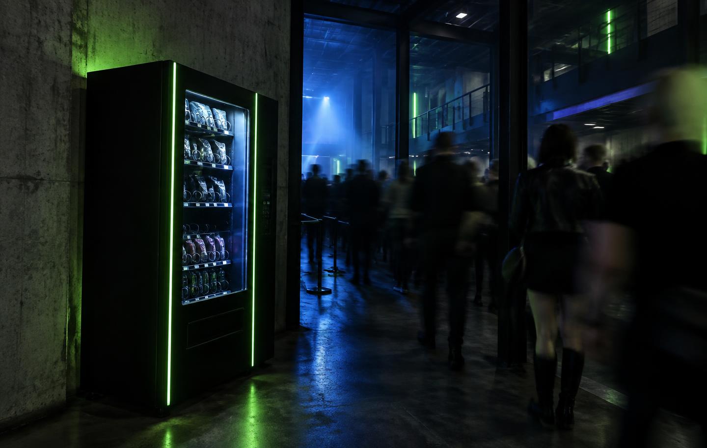 A vendo lol machine lit in a dark venue corridor while people walk past